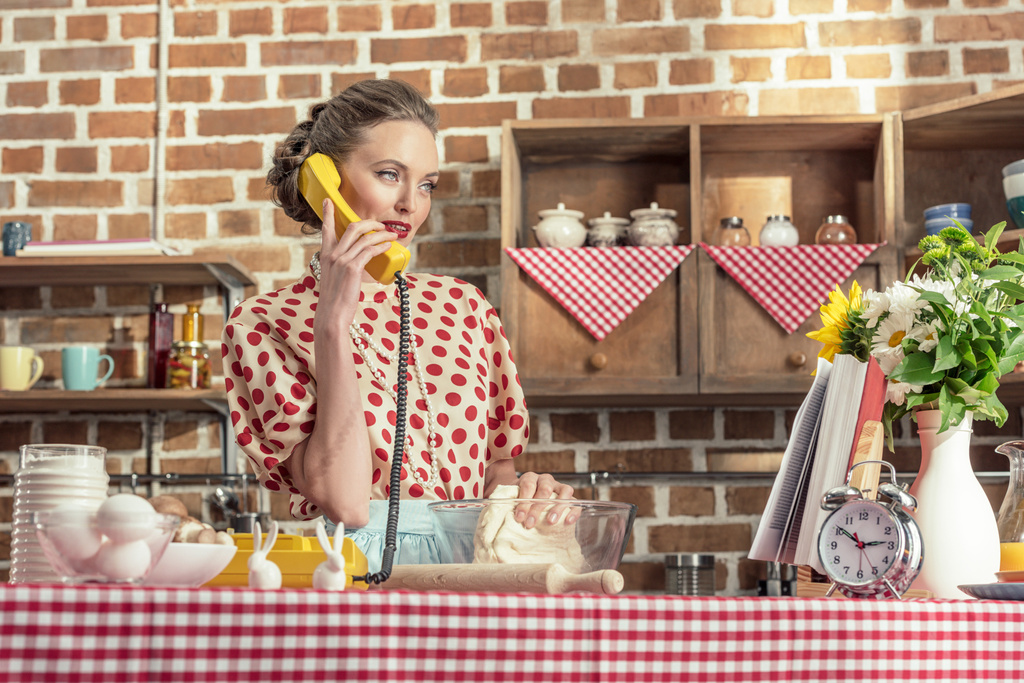όμορφη νοικοκυρά ενηλίκων μιλώντας από τηλεφώνου και ζύμωμα ζύμη στην κουζίνα - Φωτογραφία, εικόνα