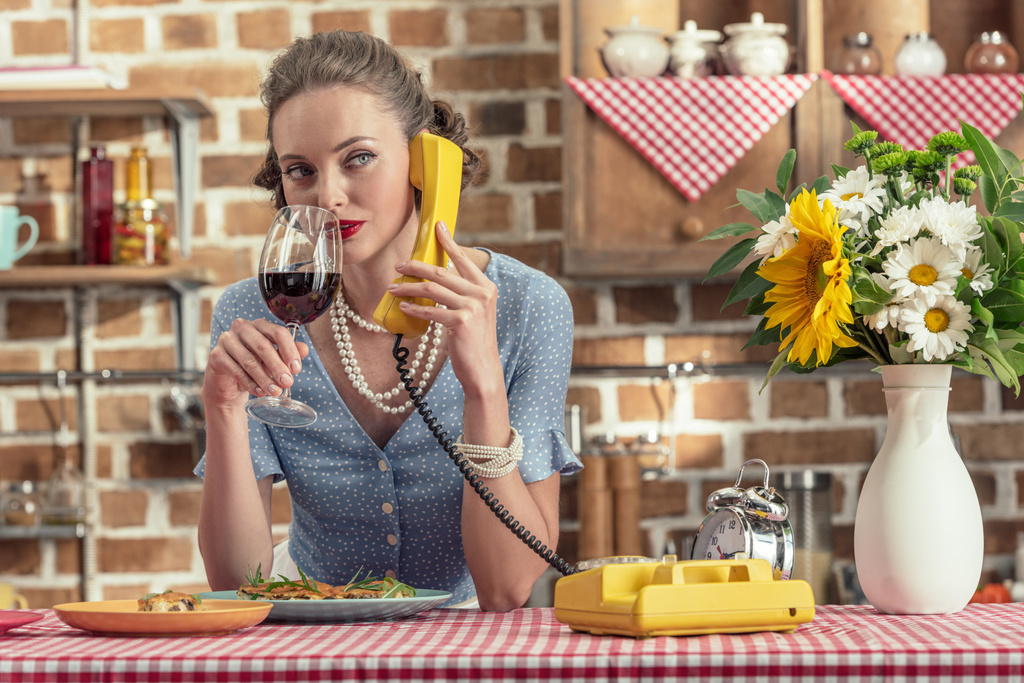 bela dona de casa adulta com copo de vinho falando por telefone rotativo vintage na cozinha
 - Foto, Imagem