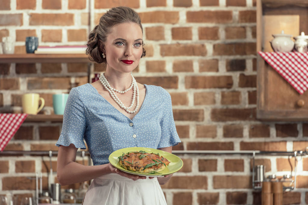 smiling adult housewife in vintage clothes holding plate with mushroom cake at kitchen - Photo, Image