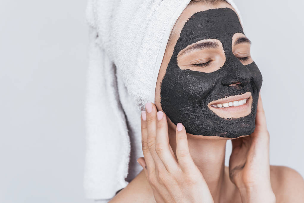 Close-up shot van jonge mooie vrouwelijke zwarte klei masker, toe te passen schoonheid Wellness-behandeling op haar gezicht huid doen. Horizontale schot van vrouwelijke toepassen organische gezichts masker, met gesloten ogen op wit - Foto, afbeelding