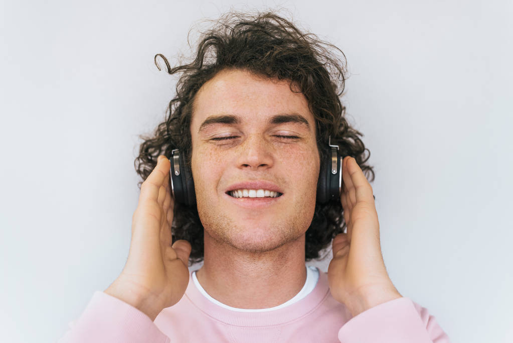 Detailní portrét pohledný usmívající se mladý mužský model s kudrnatými vlasy a pihy na sobě růžové šaty, poslech hudby ve sluchátkách, izolované na bílém studiu zdi. Kopírovat prostor pro reklamu - Fotografie, Obrázek