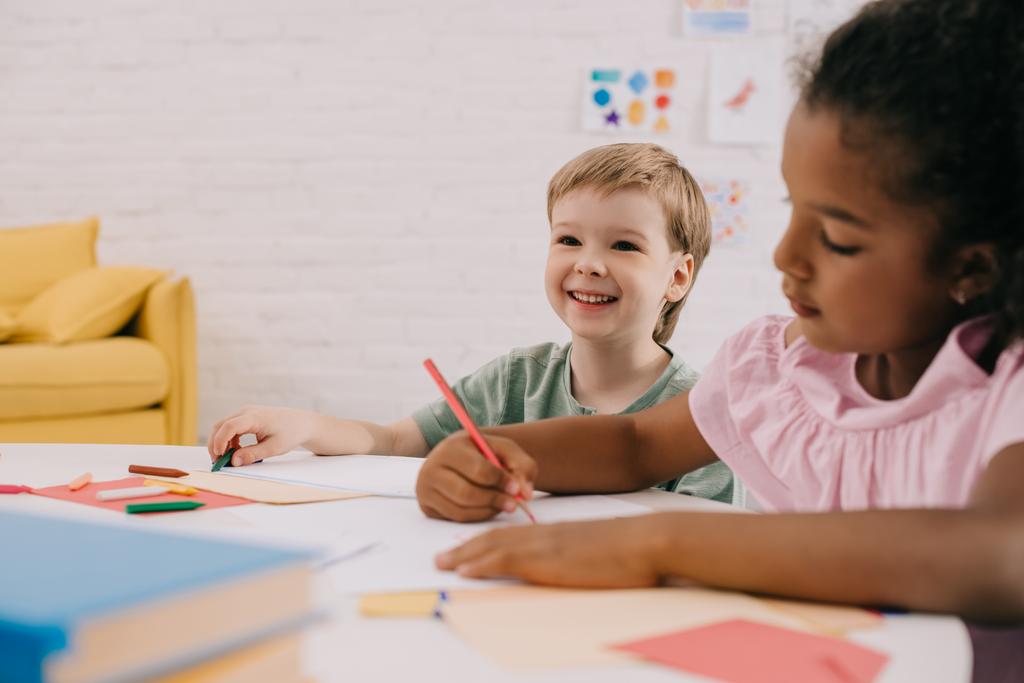 focus selettivo di bambini in età prescolare multiculturale a tavola con carte e matite in classe
 - Foto, immagini
