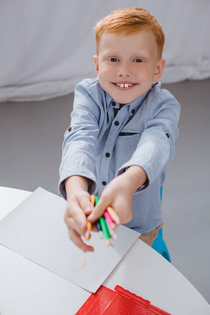 веселий дошкільник червоне волосся хлопчик з олівцями сидить за столом з папером для малювання в класі
 - Фото, зображення