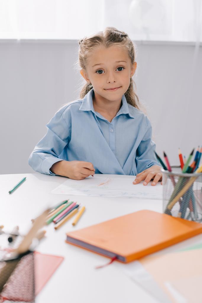 πορτρέτο του χαμογελαστό παιδί που κάθεται στο τραπέζι με πολύχρωμα μολύβια και έγγραφα για την κατάρτιση - Φωτογραφία, εικόνα