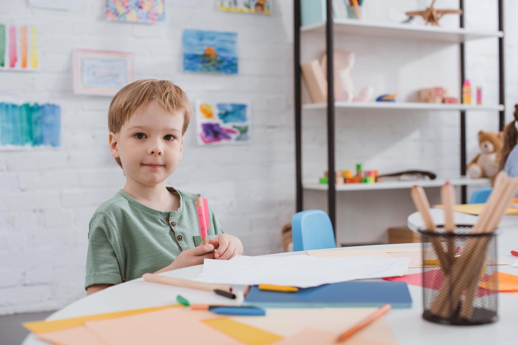 portrait of preschooler boy sitting at table with paper and colorful pencils for drawing in classroom - Photo, Image