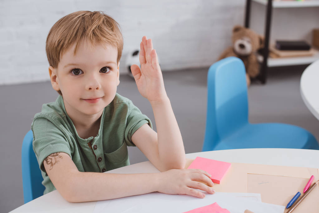 портрет милого дошкольника с поднятой рукой, сидящего за столом в классе
 - Фото, изображение