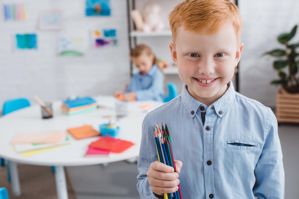 selective focus of happy red hair boy with pencils and classmate at table in classroom  - Photo, Image