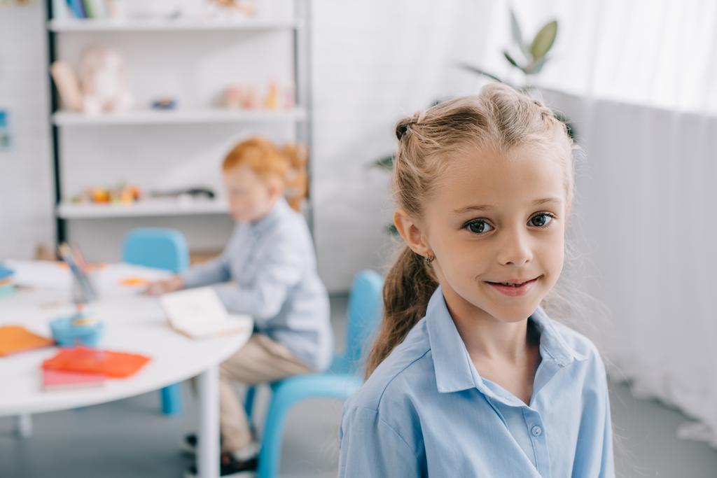 Selektiver Fokus eines lächelnden Vorschülers, der in die Kamera blickt, mit einem Mitschüler am Tisch im Klassenzimmer  - Foto, Bild