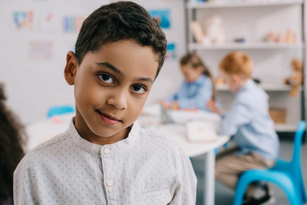 szelektív összpontosít, aranyos afro-amerikai fiú nézte kamera osztálytársai mögött a tanterem - Fotó, kép