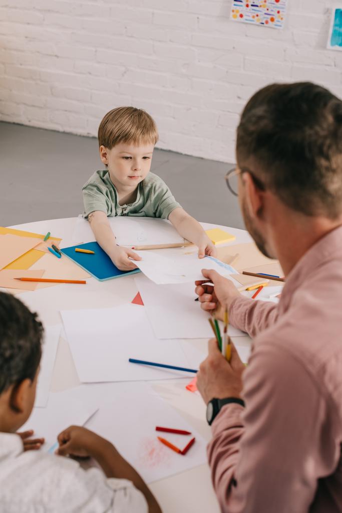 enfoque selectivo del maestro y los niños multirraciales dibujo de dibujos con lápices de colores en la mesa en el aula
 - Foto, Imagen