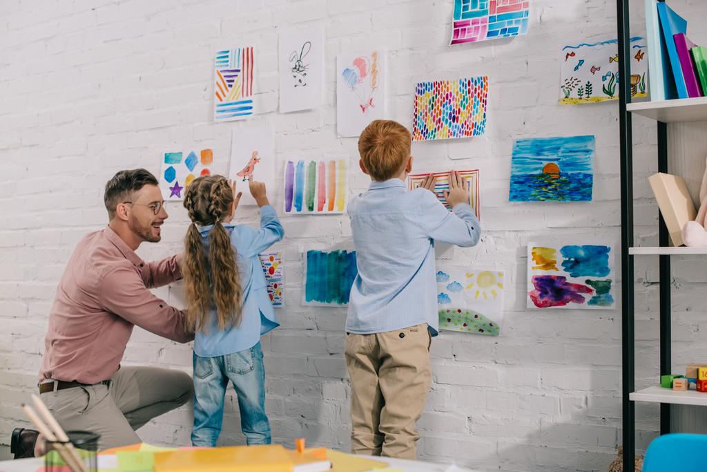 Улыбающийся учитель помогает маленьким дошкольникам повесить красочные картинки на стену в классе
 - Фото, изображение