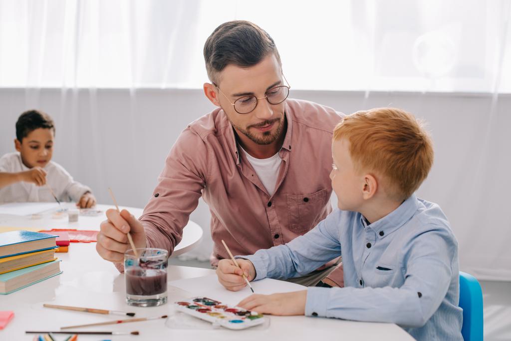 Lehrerin hilft kleinem Jungen beim Malen am Tisch im Klassenzimmer - Foto, Bild