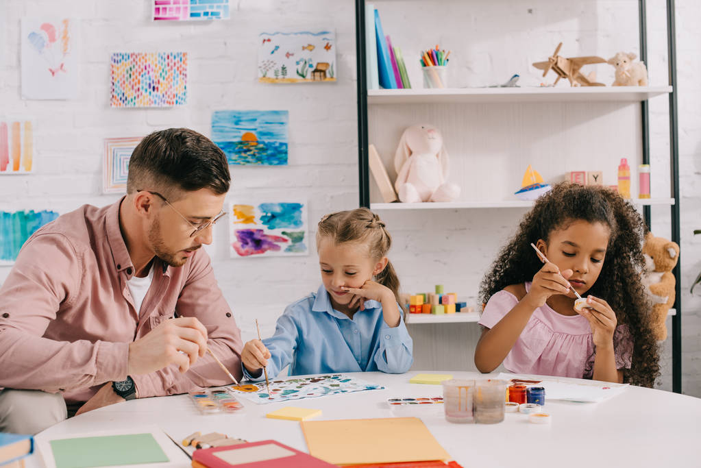 δάσκαλος σε γυαλιά οράσεως και πολυφυλετική παιδιά αντλώντας εικόνες με χρώματα στο τραπέζι στην τάξη - Φωτογραφία, εικόνα
