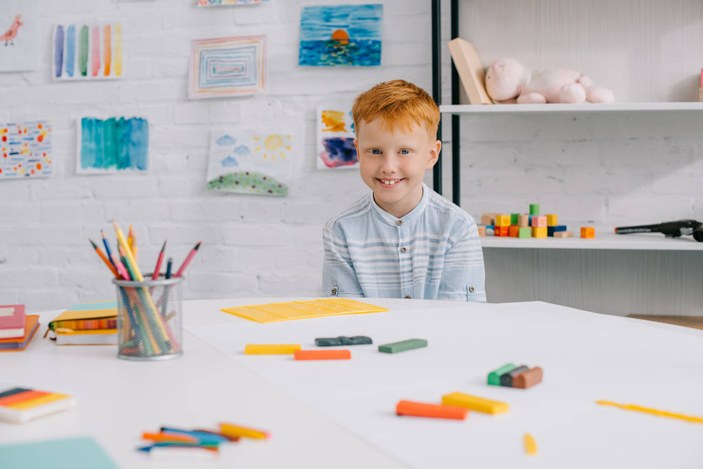 Porträt eines lächelnden Jungen mit rotem Haar, der am Tisch sitzt und bunte Knetmasse für die Bildhauerei im Raum verwendet - Foto, Bild