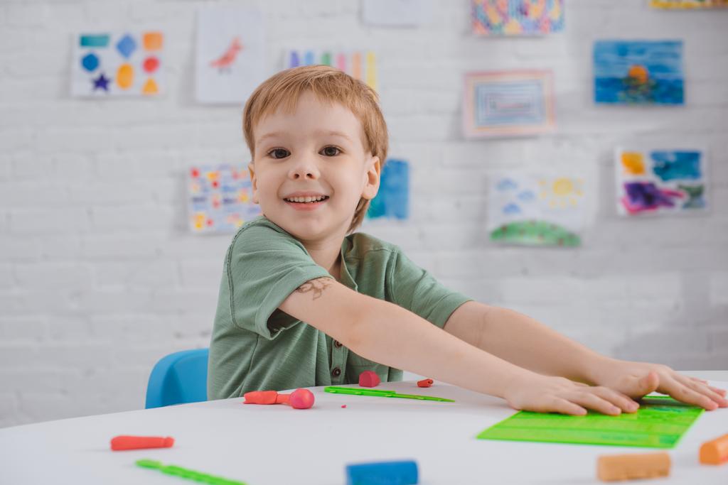 портрет усміхненого хлопчика, що сидить за столом з барвистим пластиліном для скульптури в кімнаті
 - Фото, зображення