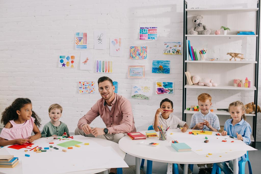 uśmiechający się nauczycielka i wieloetnicznym dzieci siedzi przy stole z plasteliny kolorowe w klasie - Zdjęcie, obraz