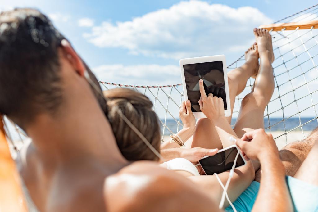 デジタル デバイスを使用して、海の近くのビーチのハンモックでリラックスしながら若いカップル - 写真・画像
