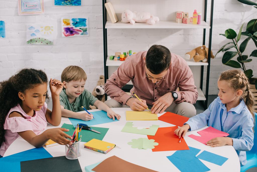 enfants d'âge préscolaire multiethnique et enseignants faisant appliquer du papier à table en classe
 - Photo, image