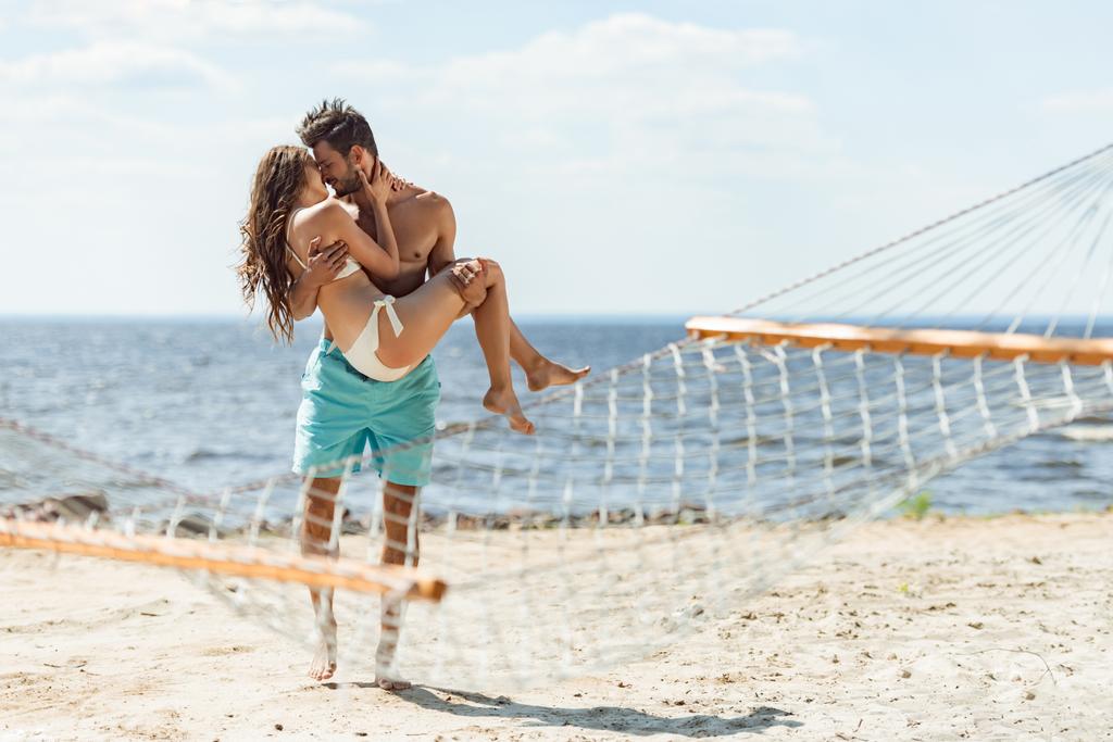 мужчина держит девушку на руках и целует ее на пляже, с гамаком на переднем плане
 - Фото, изображение