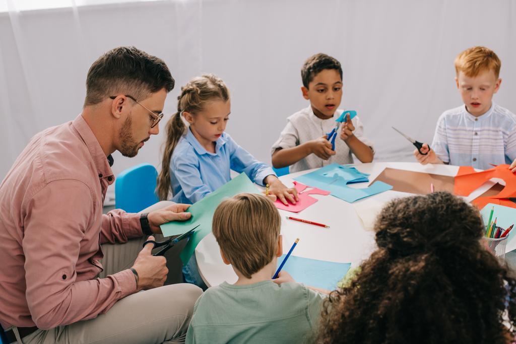 vista parcial del maestro masculino y niños preescolares multirraciales cortando papeles coloridos con tijeras en la mesa en el aula
 - Foto, Imagen
