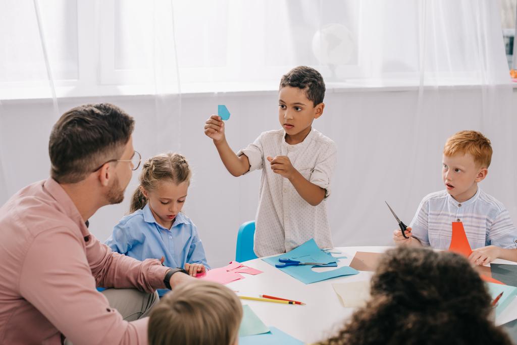 gedeeltelijke weergave van mannelijke leraar en multiraciale kleuters zitten aan tafel met kleurrijke papieren in klas - Foto, afbeelding