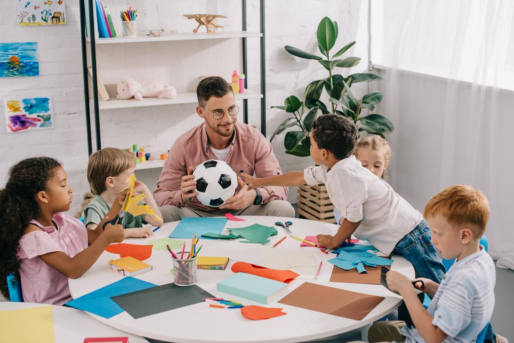 muž učitel s fotbalovým míčem a mnohonárodnostní předškolní děti sedí u stolu s barevné papíry v učebně - Fotografie, Obrázek