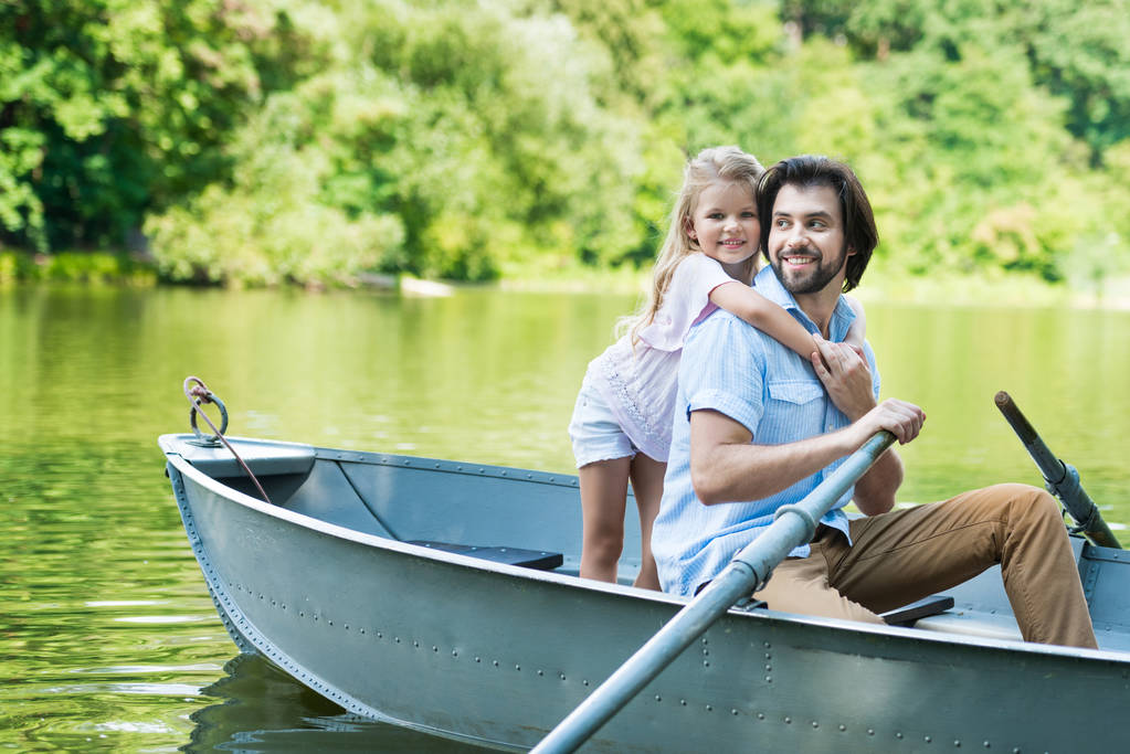 счастливая дочь обнимает отца сзади, пока они катаются на лодке по озеру в парке
 - Фото, изображение