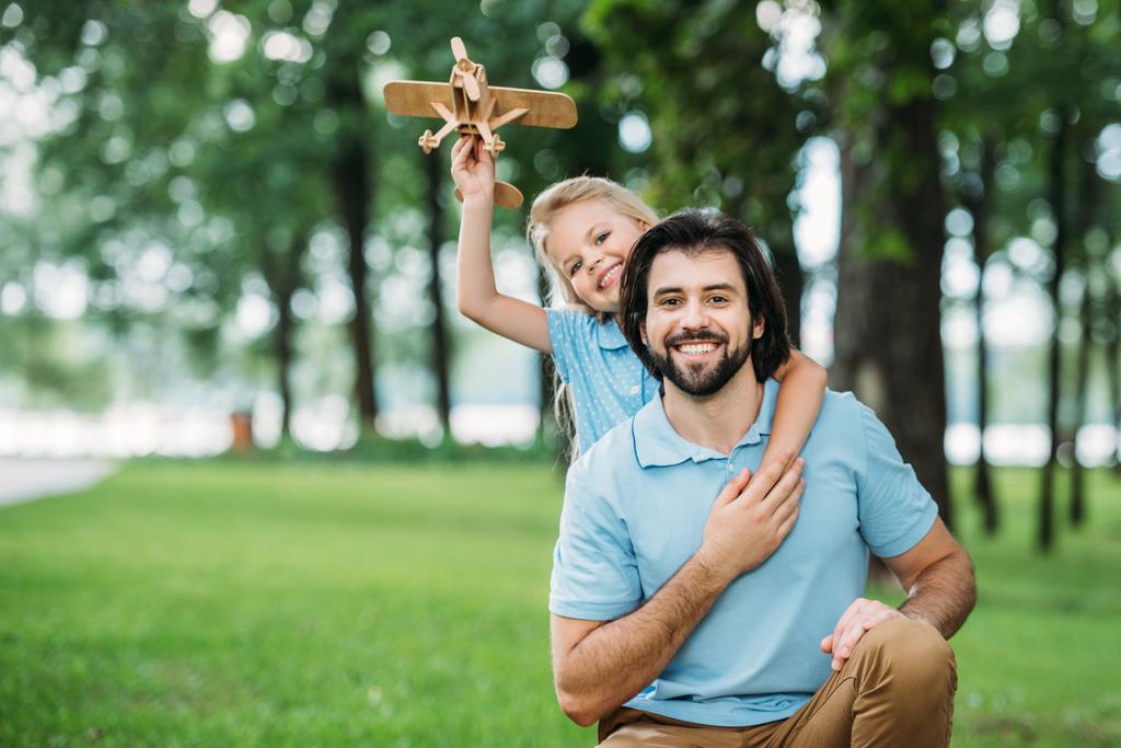 Entzückend lächelnde Tochter umarmt glücklichen Vater von hinten und hält Spielzeugflugzeug im Park - Foto, Bild