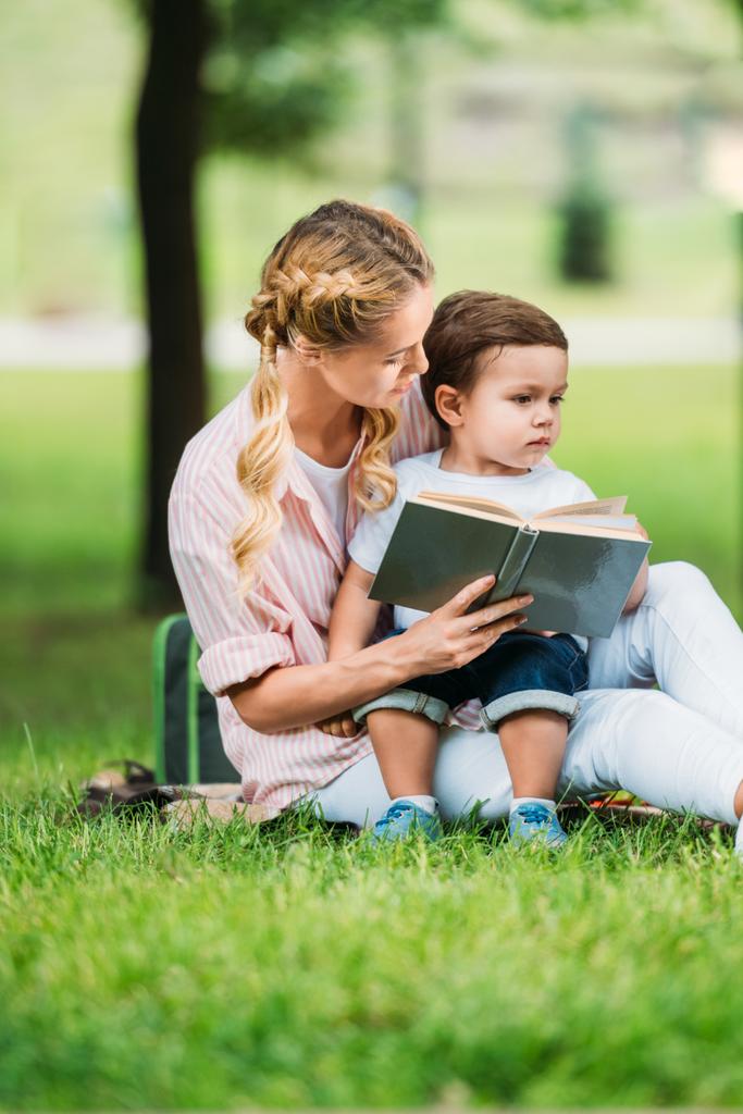 mother and son sitting with book on grass at park - Photo, Image