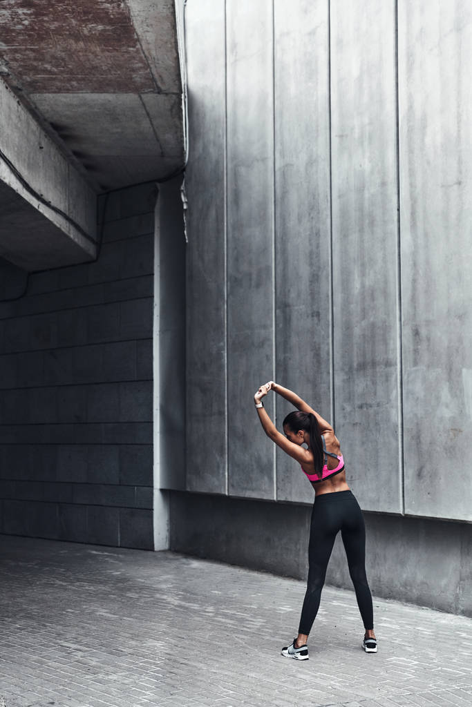 back view of sportive muscular woman in pink top stretching outdoors at grey wall  - Photo, Image