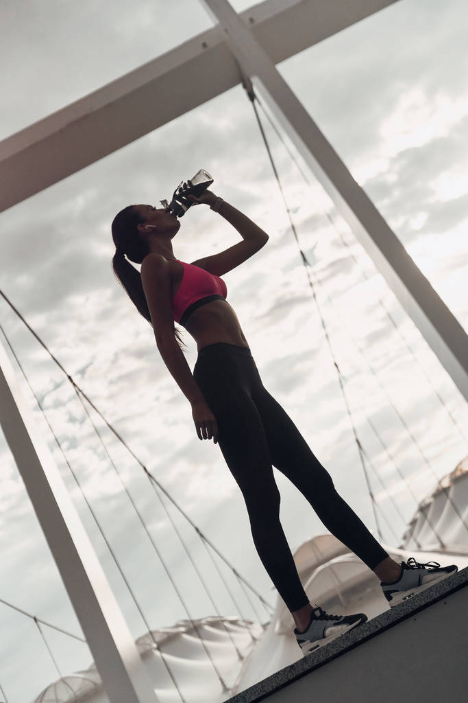 janoinen nainen urheiluvaatteet juomaveden muodossa pullon seisoessaan ulkona
 - Valokuva, kuva
