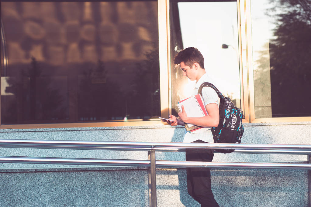 Φοιτητής χρησιμοποιώντας το κινητό τηλέφωνο, κρατώντας ένα τετράδιο και μεταφέρουν ένα σακίδιο με τα πόδια μπροστά από το κτήριο του Πανεπιστημίου. Νεαρό αγόρι που φοράει μπλε πουκάμισο και σκούρο τζιν - Φωτογραφία, εικόνα