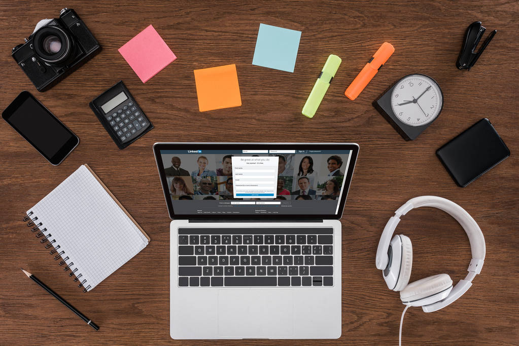 Draufsicht auf Holztisch mit leerem Lehrbuch, Smartphone, Fotokamera und Laptop mit Linkedin auf dem Bildschirm - Foto, Bild