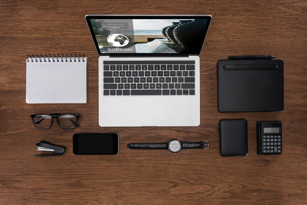 Ansicht des Arbeitsplatzes mit geordnetem leeren Lehrbuch, Smartphone, Armbanduhr und Laptop mit Buchung auf dem Bildschirm - Foto, Bild