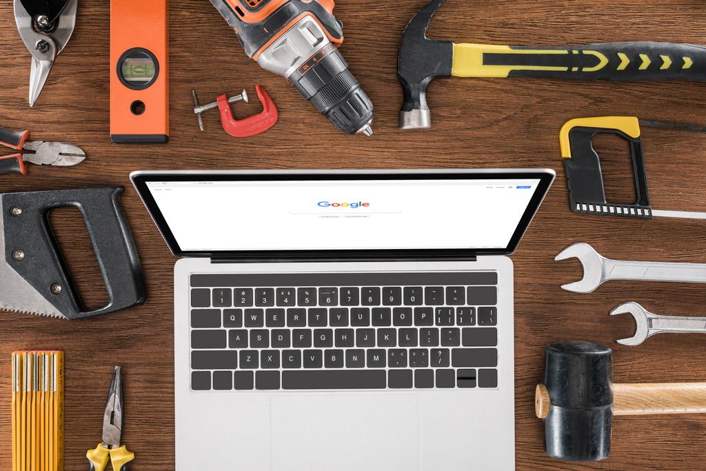 Ansicht des Arbeitsplatzes mit Laptop mit Google auf dem Bildschirm, umgeben von verschiedenen Werkzeugen auf Holztisch  - Foto, Bild