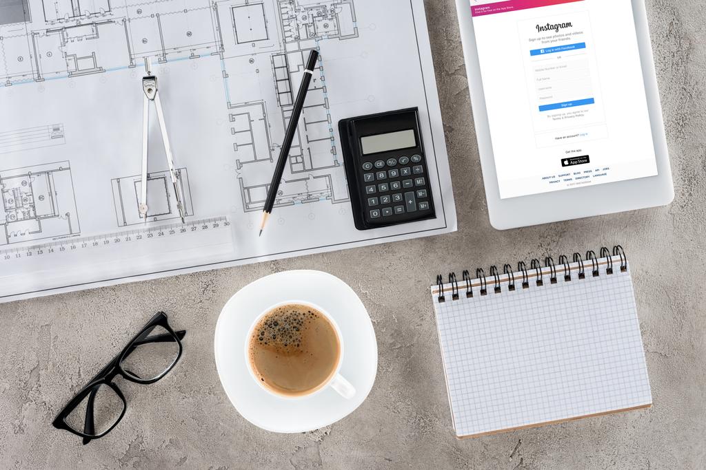 Ansicht des Architekten-Arbeitsplatzes mit Blaupause, Trennwand, Kaffee und digitalem Tablet mit Instagram auf dem Bildschirm - Foto, Bild