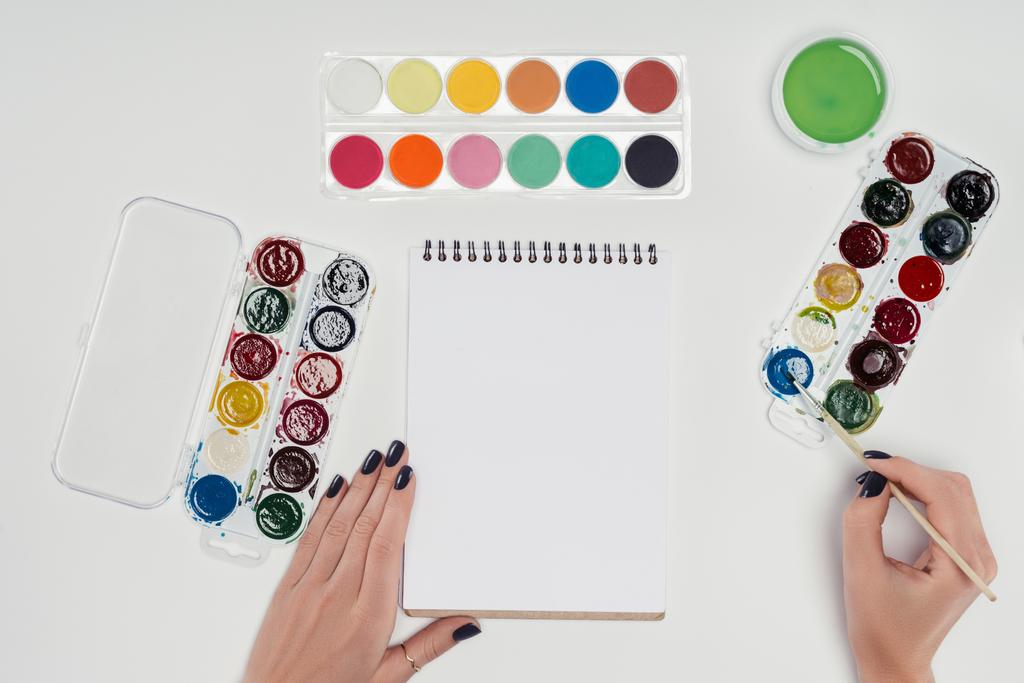 обрезанное изображение художницы, рисующей кистью в пустом учебнике за белым столом с красочными красками
  - Фото, изображение