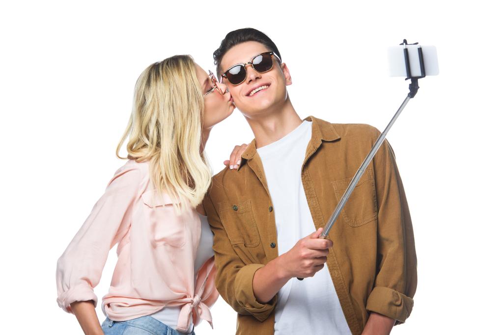 young couple taking selfie from monopod while woman kissing her boyfriend isolated on white - Photo, Image