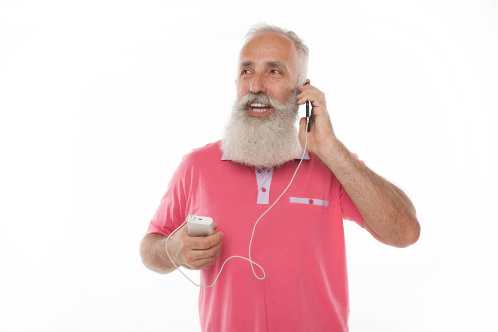 Studio shot van gelukkig senior bebaarde man die lacht tijdens het opladen en praten op mobiele telefoon tegen een witte achtergrond. Oude man opladen telefoon. PowerBank. - Foto, afbeelding