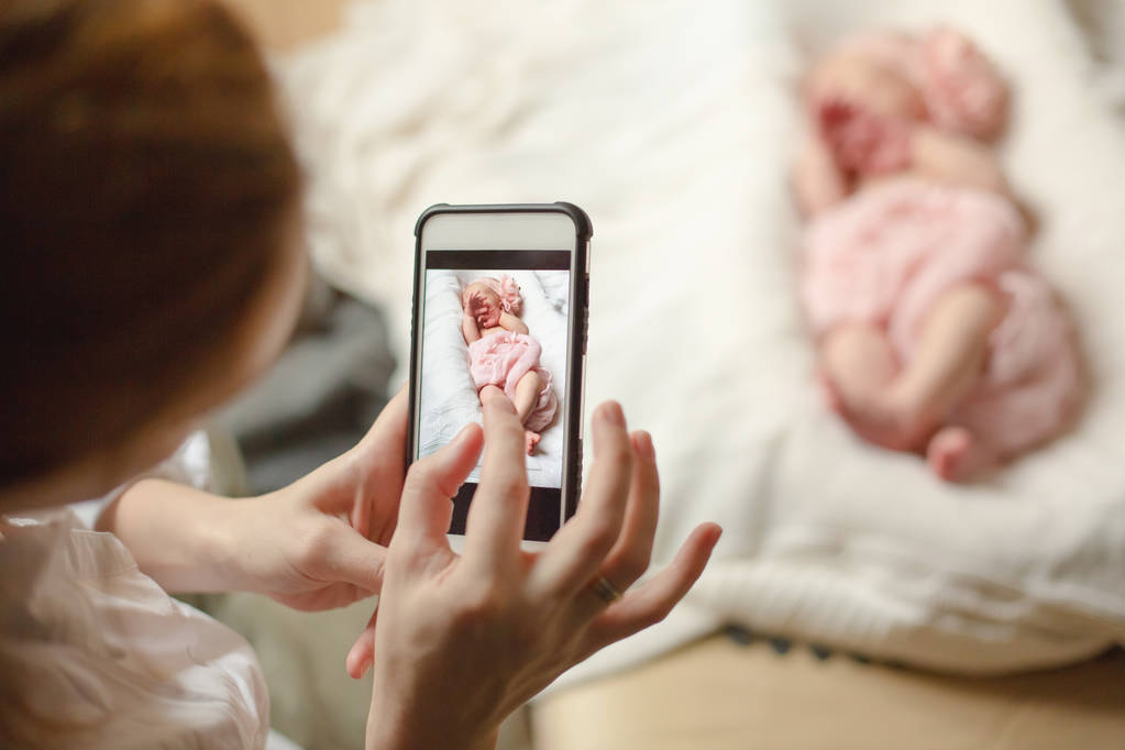 Жінка тримає смартфон і фотографує новонароджену дівчинку, яка спить на білій ковдрі. Вибірковий фокус
. - Фото, зображення