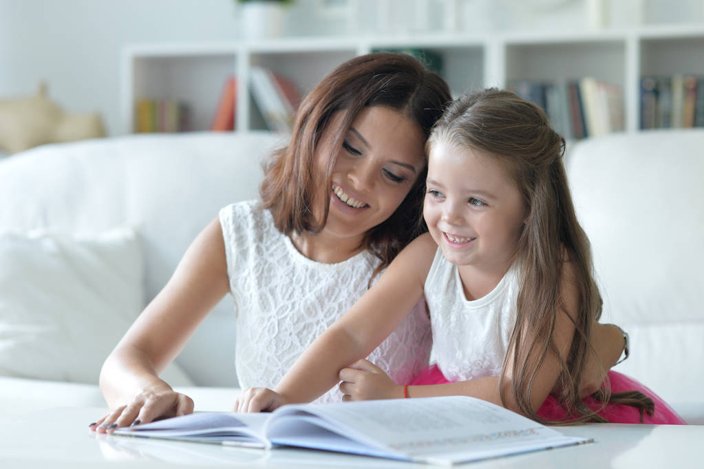 pequeña linda chica leyendo libro con madre en la mesa en casa
 - Foto, Imagen