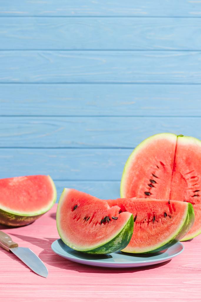 Nahaufnahme von Wassermelonenstücken und Messer auf rosa Tischplatte auf blauem Hintergrund - Foto, Bild
