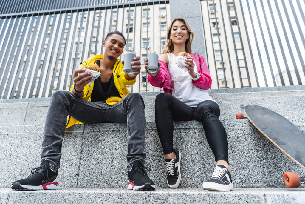 vista basso angolo di sorridente coppia multietnica di skateboarder in possesso di hamburger e mostrando bevande gassate in strada città
 - Foto, immagini