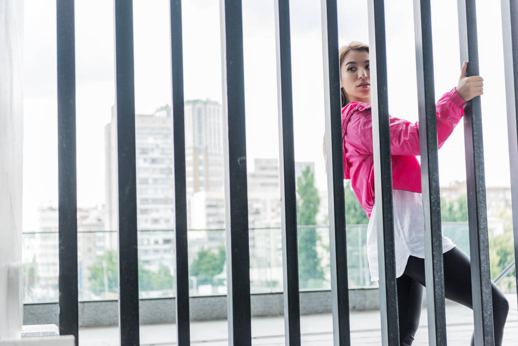 atraktivní stylová žena stojící poblíž kovový plot a koukal na městské ulici - Fotografie, Obrázek