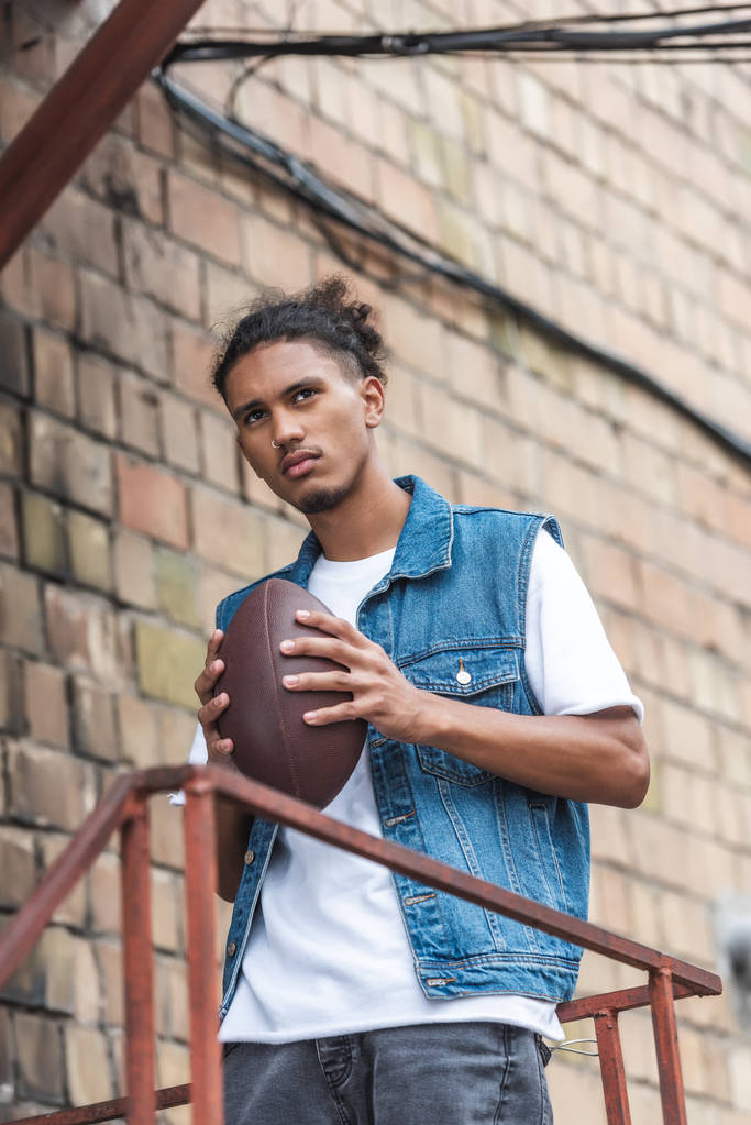 serious mixed race man holding rugby ball at urban street - Photo, Image