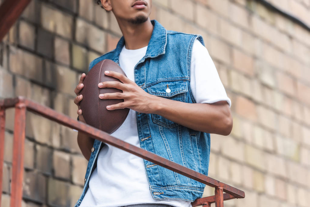 обрезанное изображение молодого человека, держащего регбийный мяч на городской улице
 - Фото, изображение