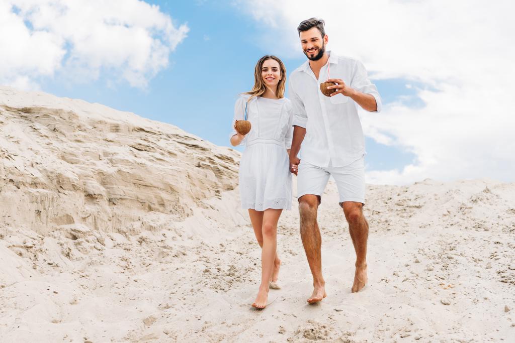 щаслива молода пара в білому з кокосовими коктейлями, що йдуть на піщаних дюнах
 - Фото, зображення