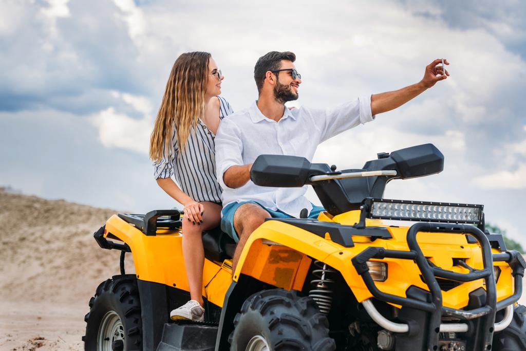jovem casal ativo tomando selfie enquanto sentado em ATV no deserto
 - Foto, Imagem