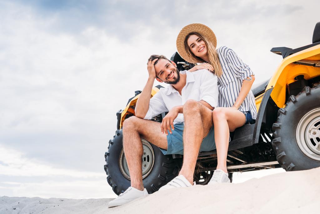 schönes junges Paar sitzt auf ATV auf Sanddüne und schaut in die Kamera - Foto, Bild