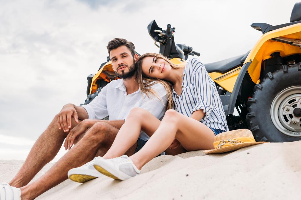 вид снизу красивой молодой пары, сидящей рядом с ATV на песчаной дюне и смотрящей в камеру
 - Фото, изображение